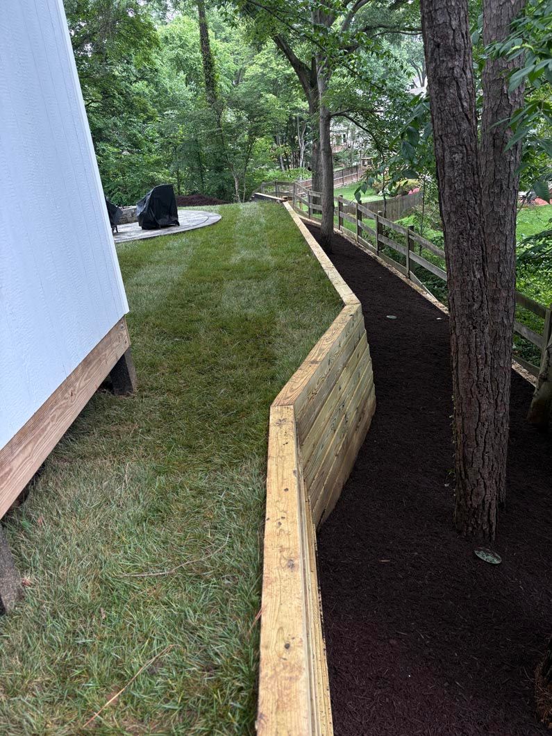 Raised grassy walkway beside a house, bordered by a wood retaining wall and trees along a dirt path