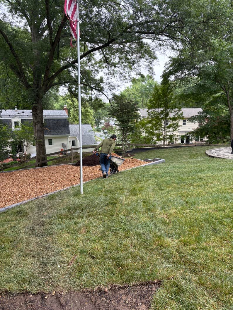 Two people kneel by a flagpole in a grassy yard with mulch, trees, and houses in the background.
