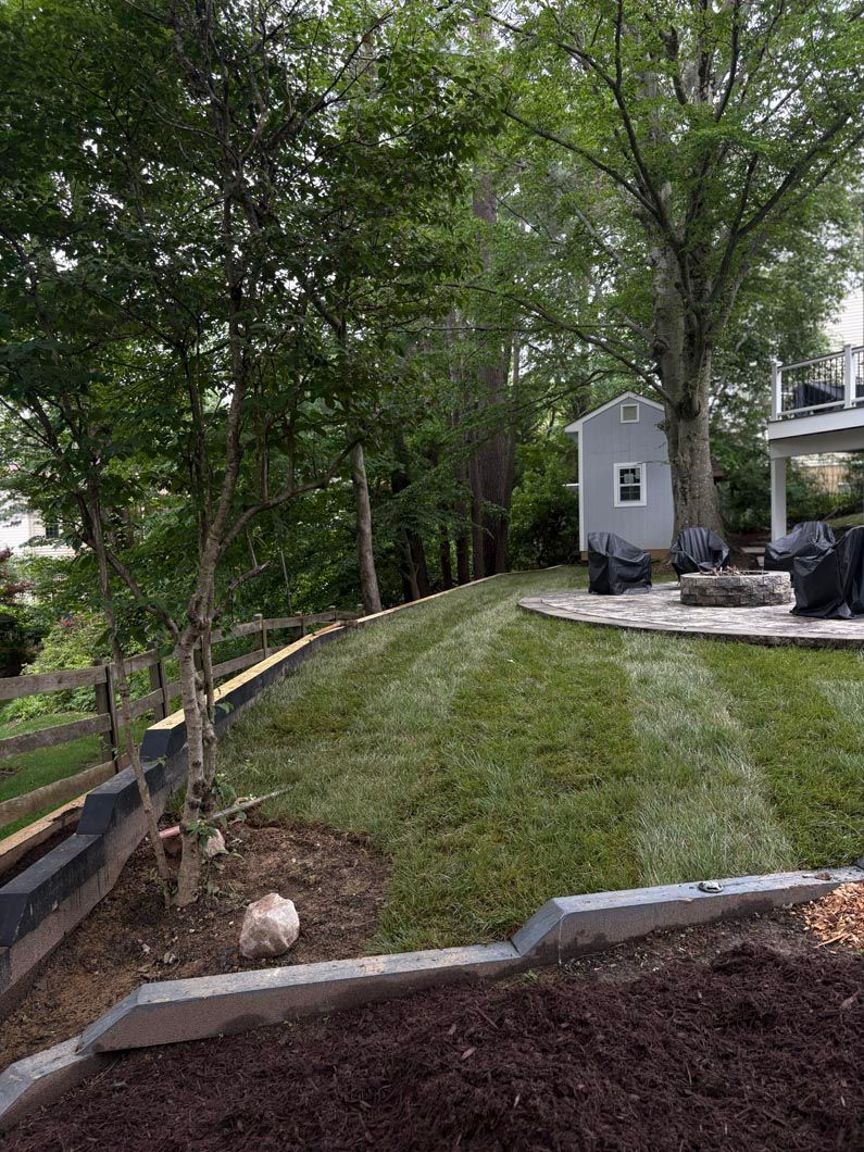 Sloped grassy yard beside a house, bordered by trees and a retaining edge with fresh mulch in front.