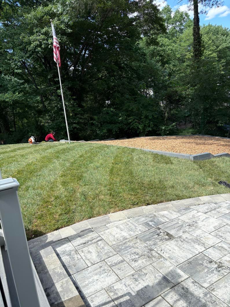 Grassy mound with a flagpole beside a stone patio, bordered by trees and a small paved path.