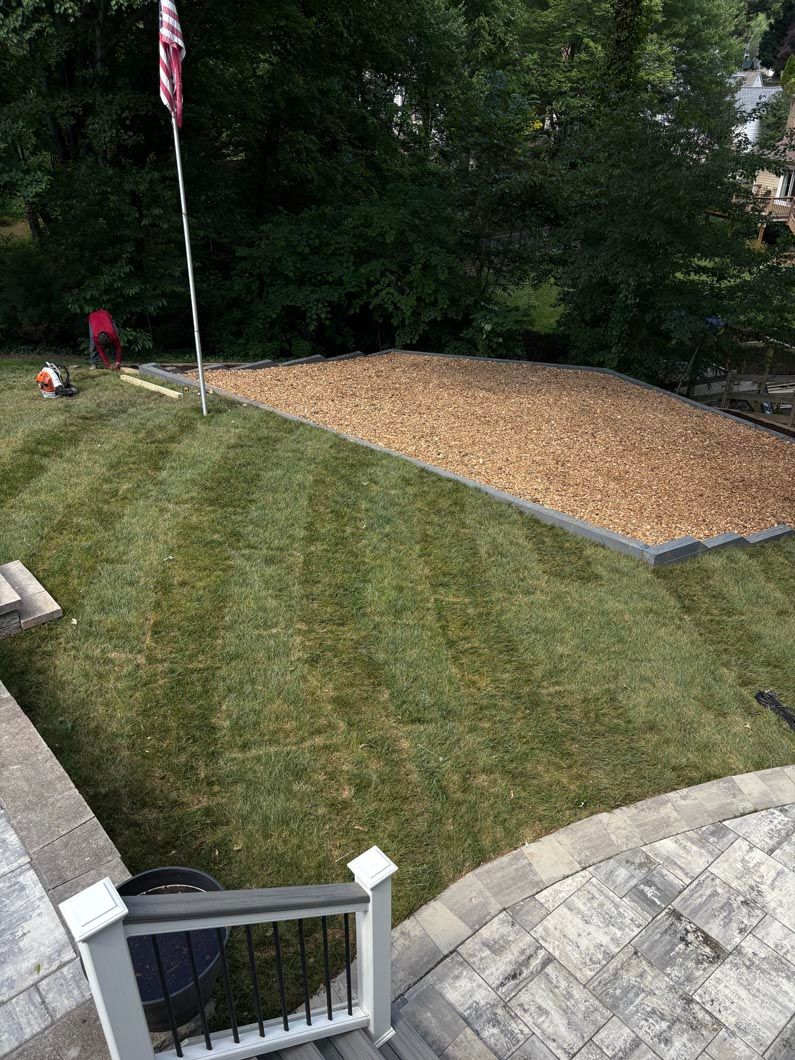 Grassy yard with a gravel driveway, stone patio, and small flagpole near trees