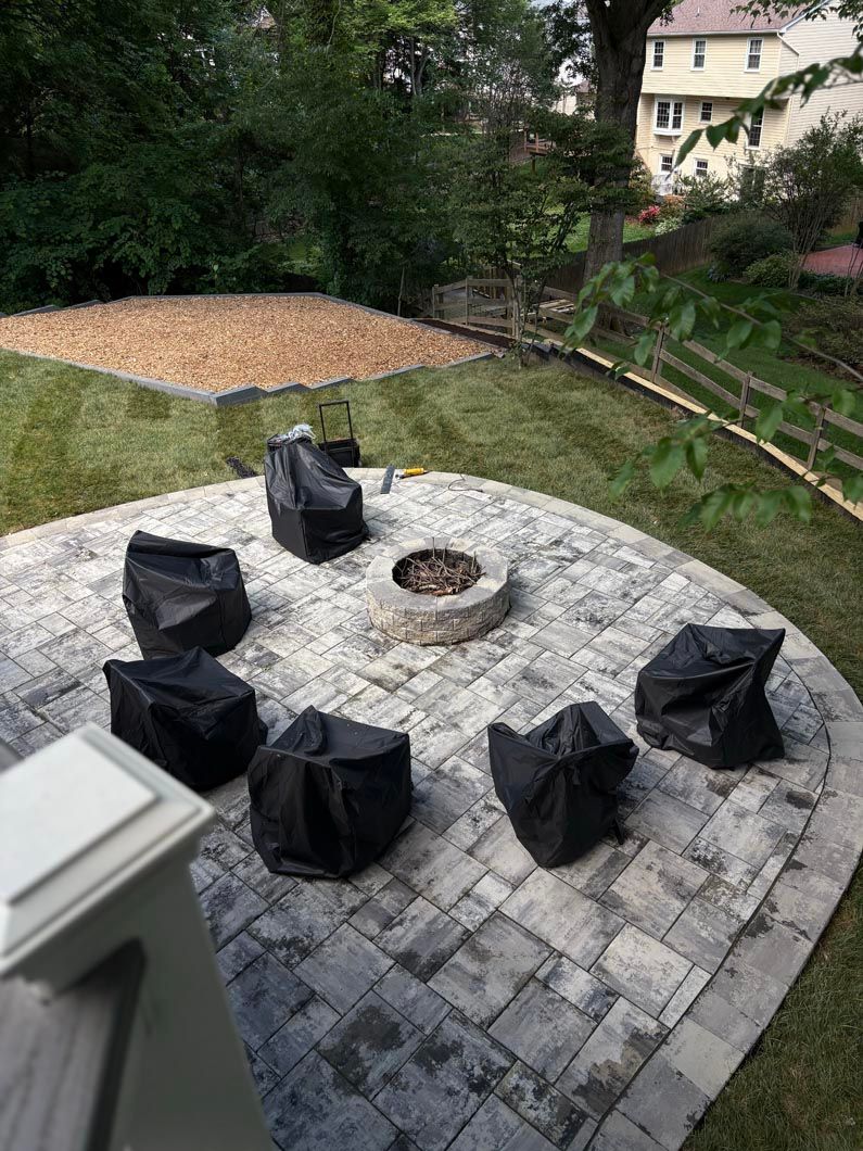 Patio with covered outdoor chairs around a fire pit, beside a grassy yard and trees