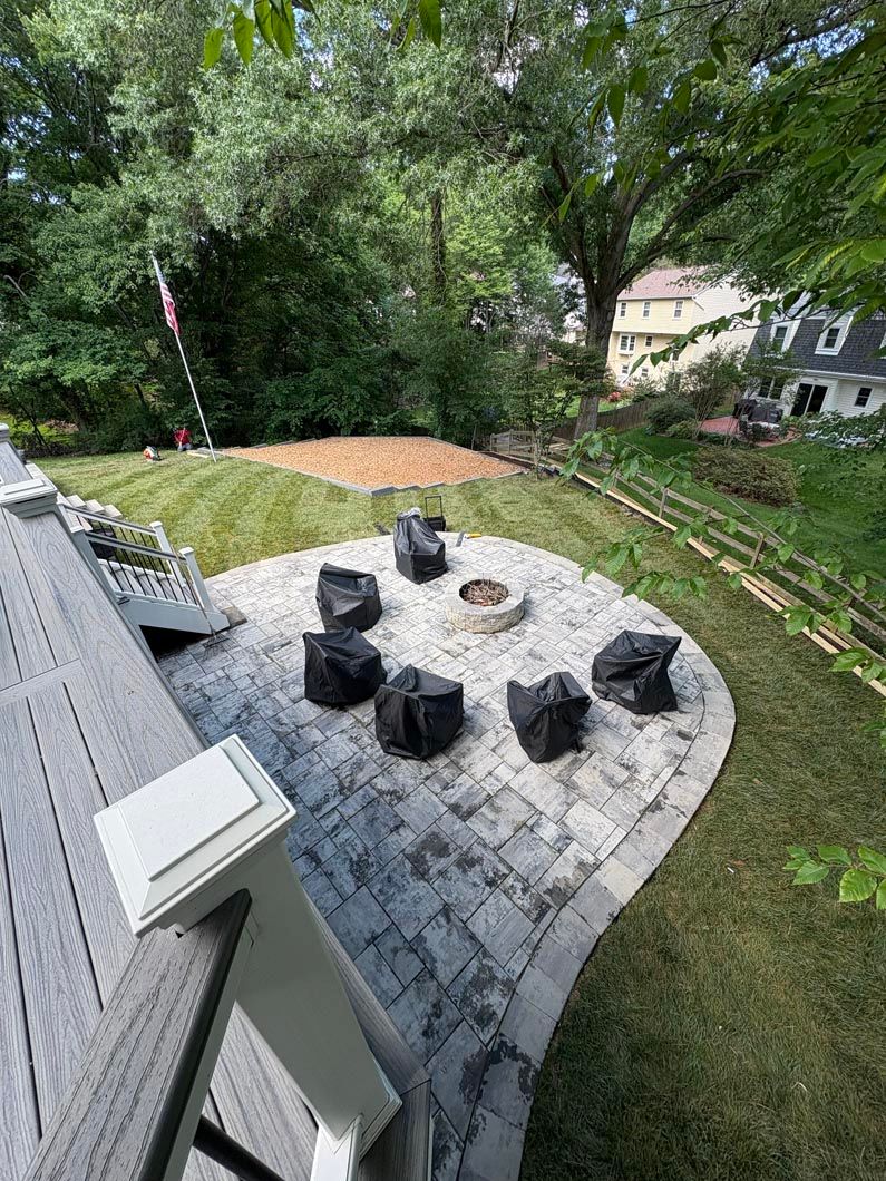 Backyard patio with six black chairs around a fire pit, bordered by green lawn and trees