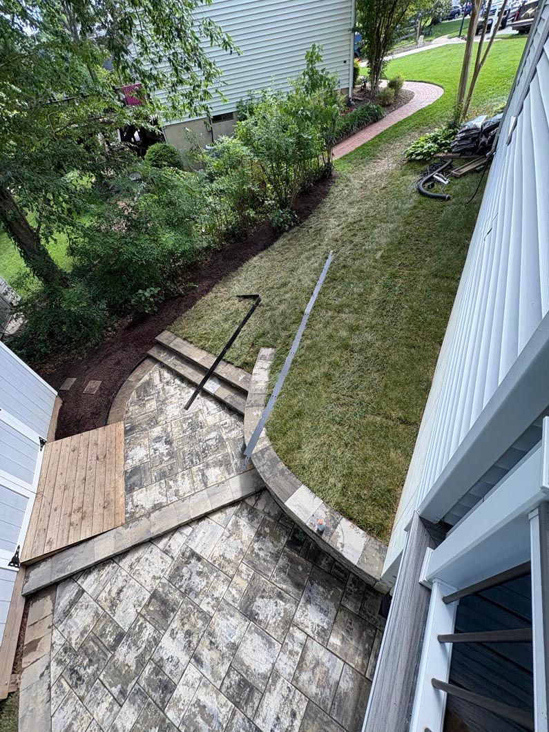 Backyard view with stone patio, grassy slope, and narrow garden path beside a house