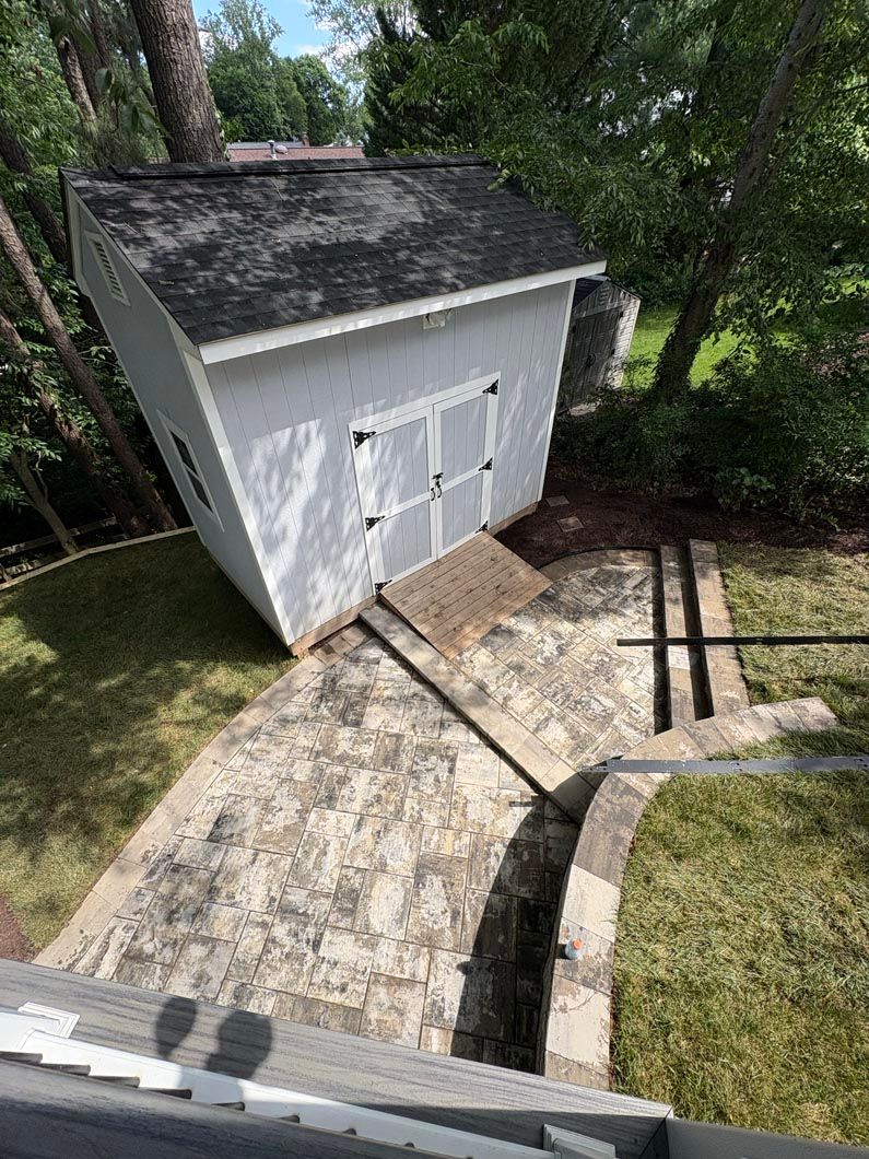 Small white shed with double doors beside a sloped concrete path and grass in a wooded yard