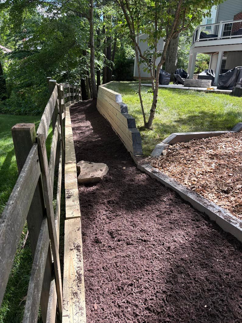 Mulched garden path beside a wooden fence and raised stone border in a leafy yard