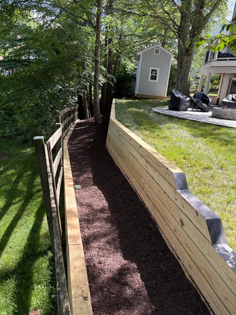 Narrow mulched walkway beside a stone retaining wall in a sunny backyard with trees and a small shed.