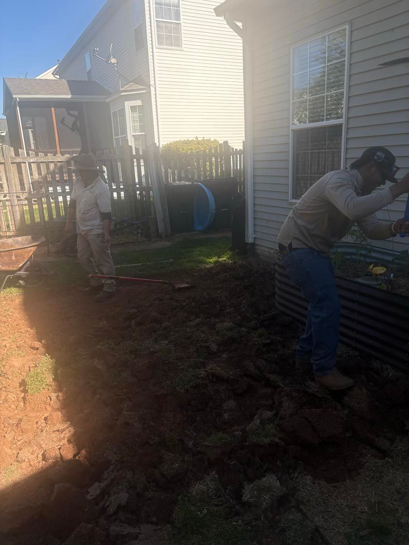 Two people work in a backyard garden beside a house, digging soil near a blue barrel.