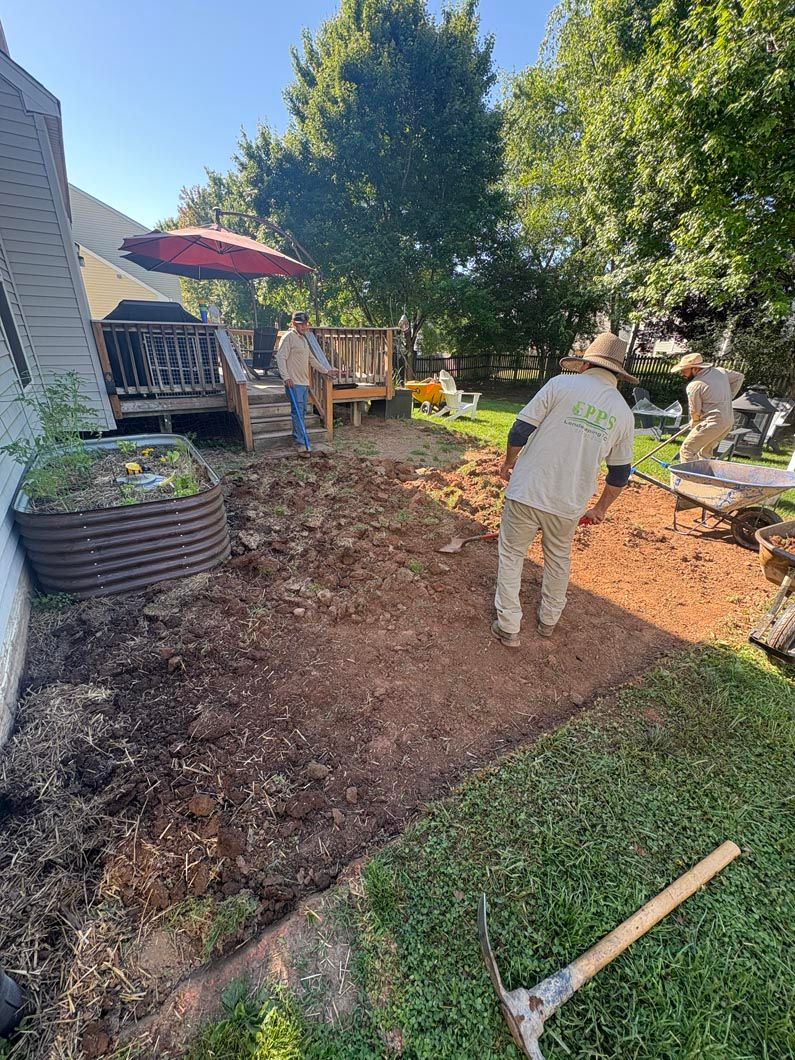 Garden workers clearing mulch and leaves in a sunny backyard beside a house.