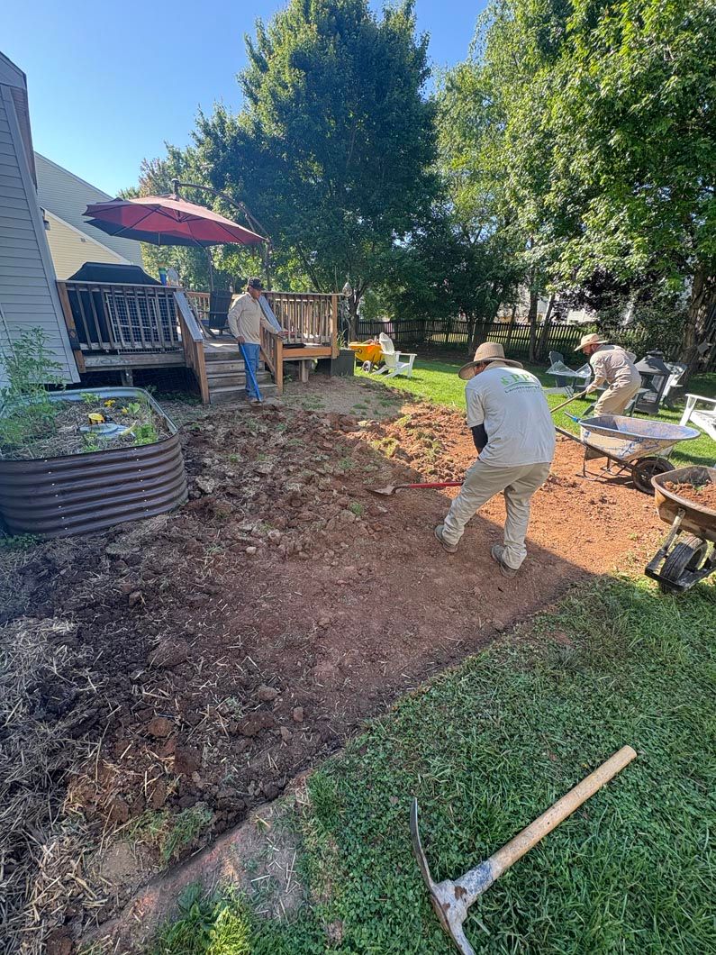 Two people landscaping a backyard with mulch, a tree, patio furniture, and a red umbrella