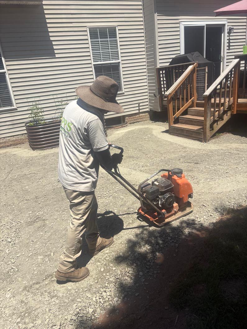 Worker using a plate compactor on gravel beside a house and porch stairs