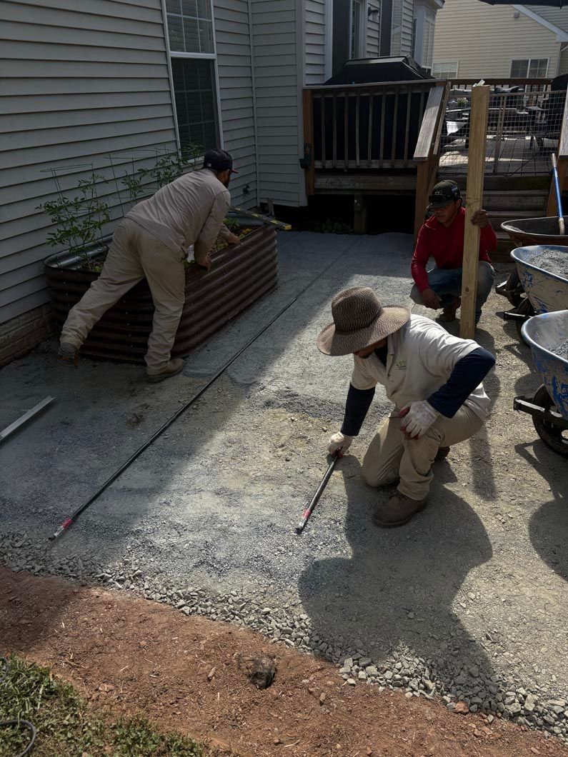 Two workers in beige coveralls spread gravel beside a house, with a wheelbarrow nearby.