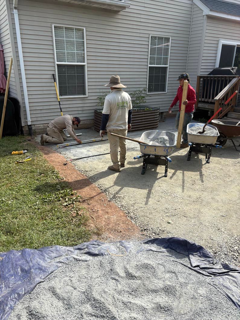 Two workers paving a backyard patio beside a house, with gravel, concrete, and wheelbarrows.