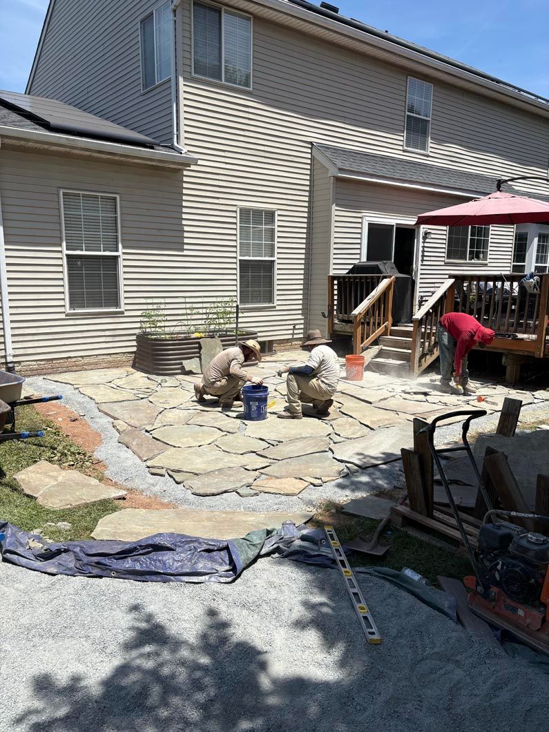Workers installing a stone patio beside a beige house, with tools and a red canopy in the yard