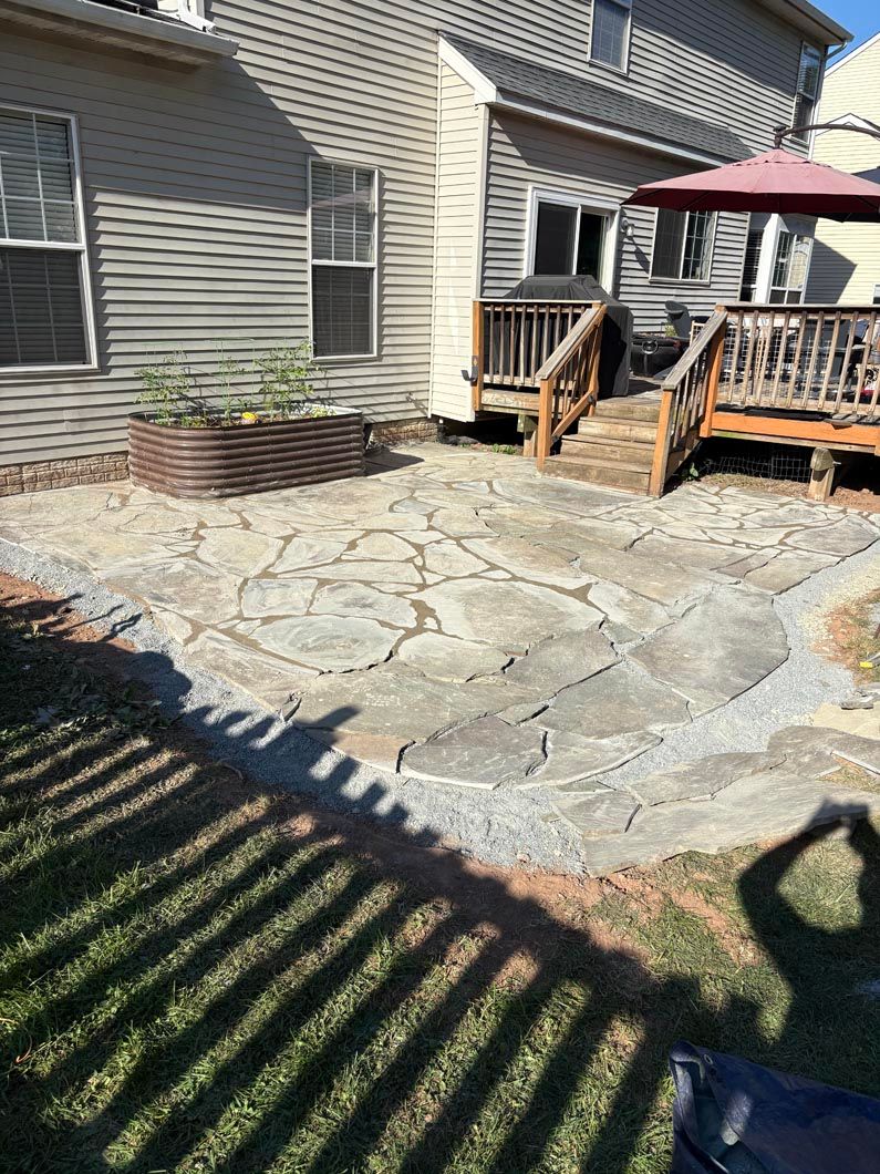 Backyard stone patio with a wooden deck, patio table with red umbrella, and raised planter by the house.