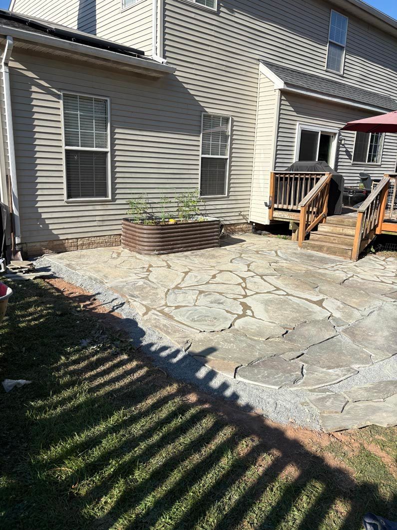 Back patio with stone pavers, small wooden deck steps, and a house exterior in sunlight