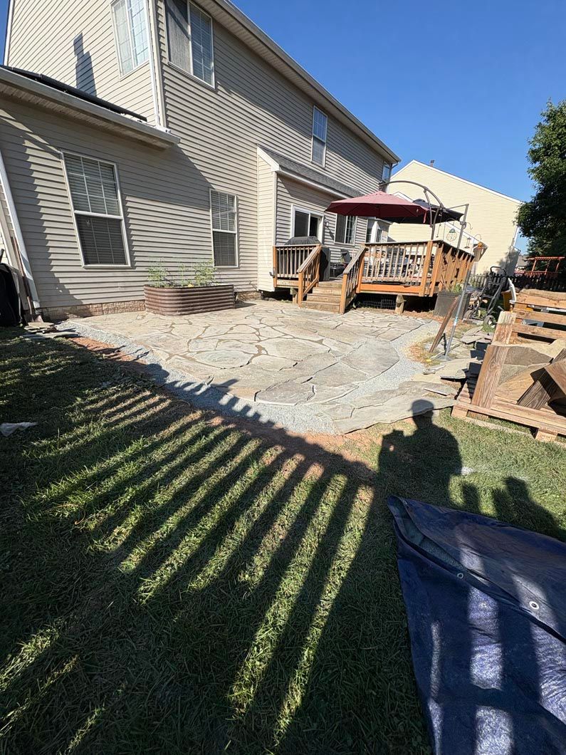 Backyard with house siding, wooden deck, and striped lawn shadows in bright sunlight