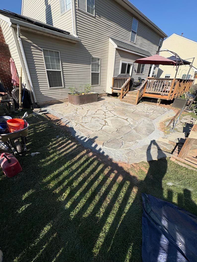 Backyard patio with stone pavers, deck chairs, a red umbrella, and house siding in bright sunlight