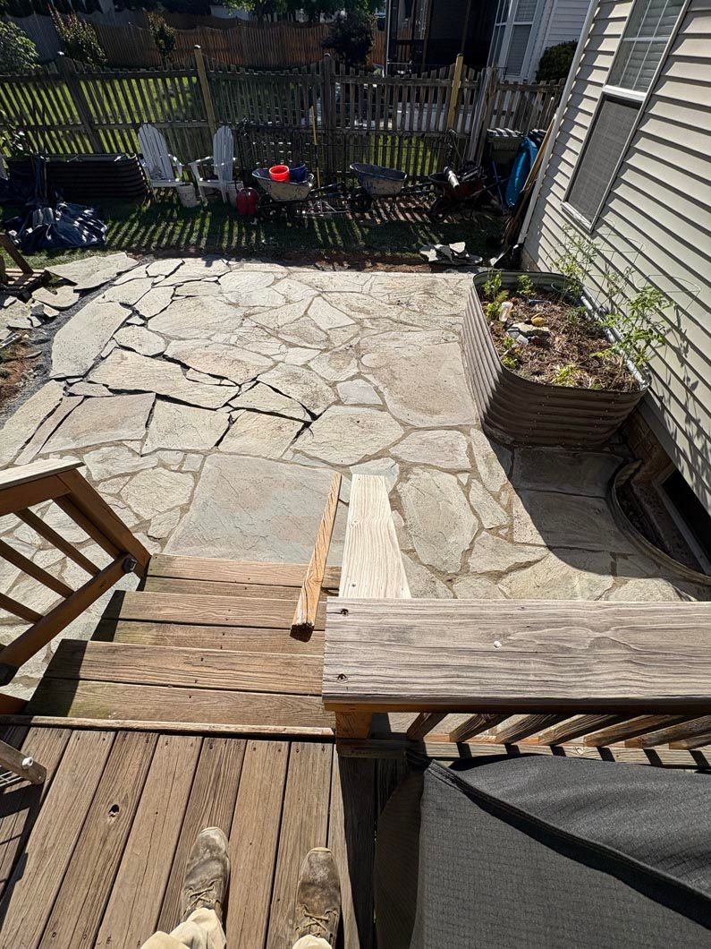 Backyard deck steps leading to a cracked stone patio with potted plants and a fence.