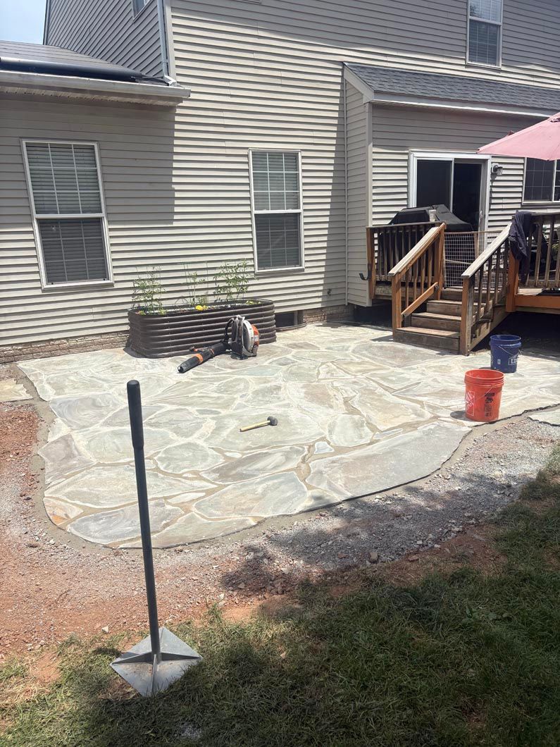 Backyard stone patio beside beige house with wooden steps and small black grill ליד.