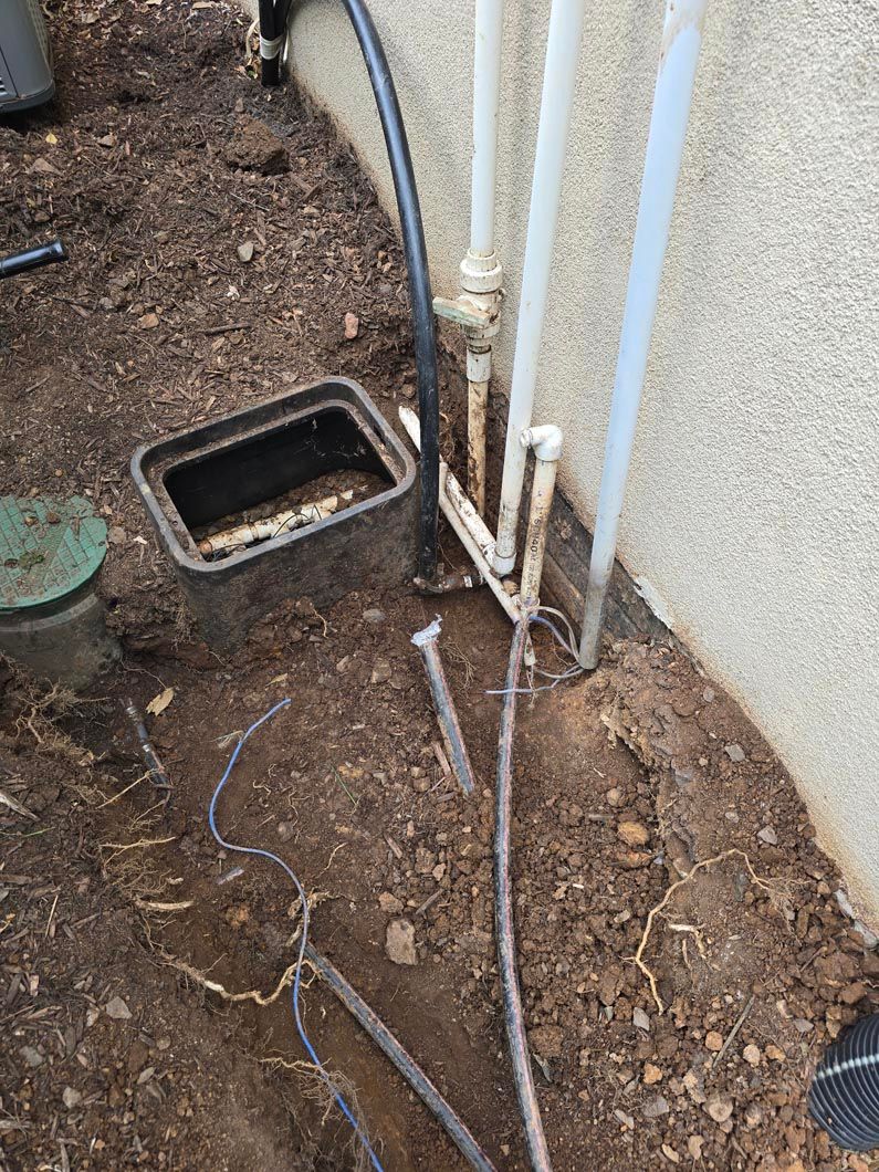 Outdoor utility pipes and a small ground access box beside a building wall with exposed cables and dirt.