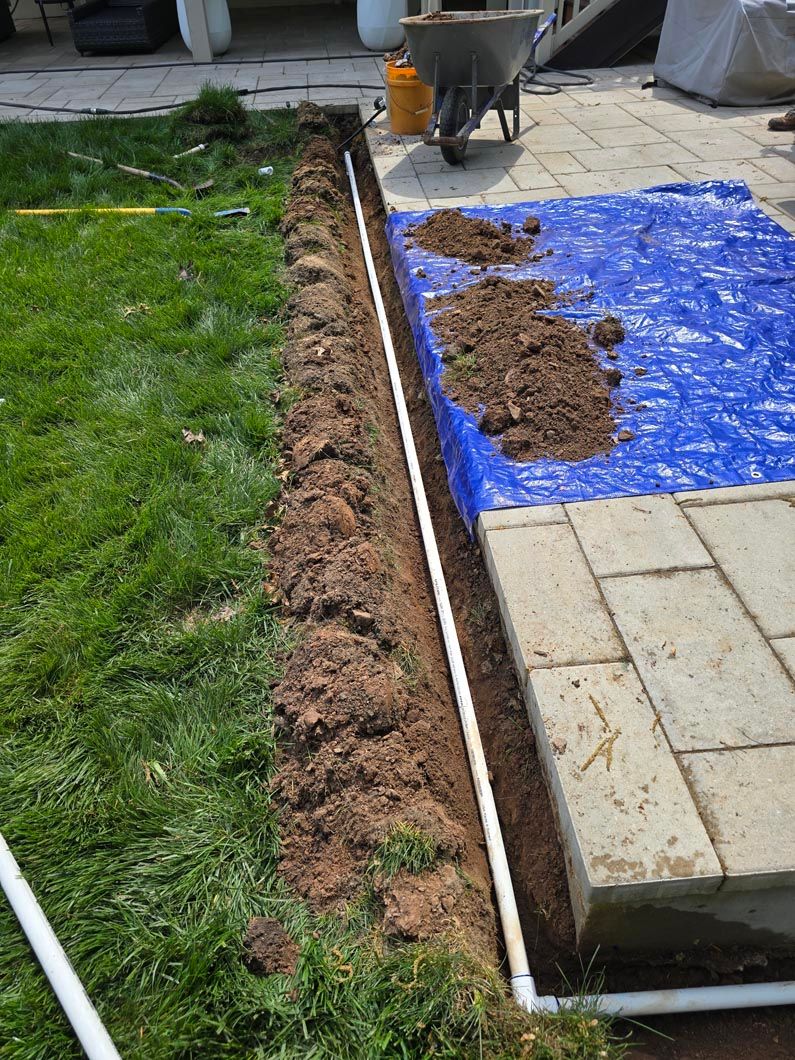 Outdoor walkway under construction beside a lawn, with soil piled on a blue tarp and edging along the path.