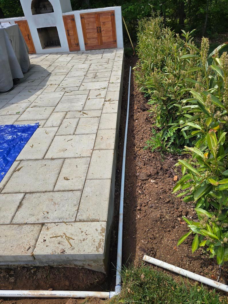 Paved patio beside a soil bed with green shrubs and a white pole; building entrance in the background.