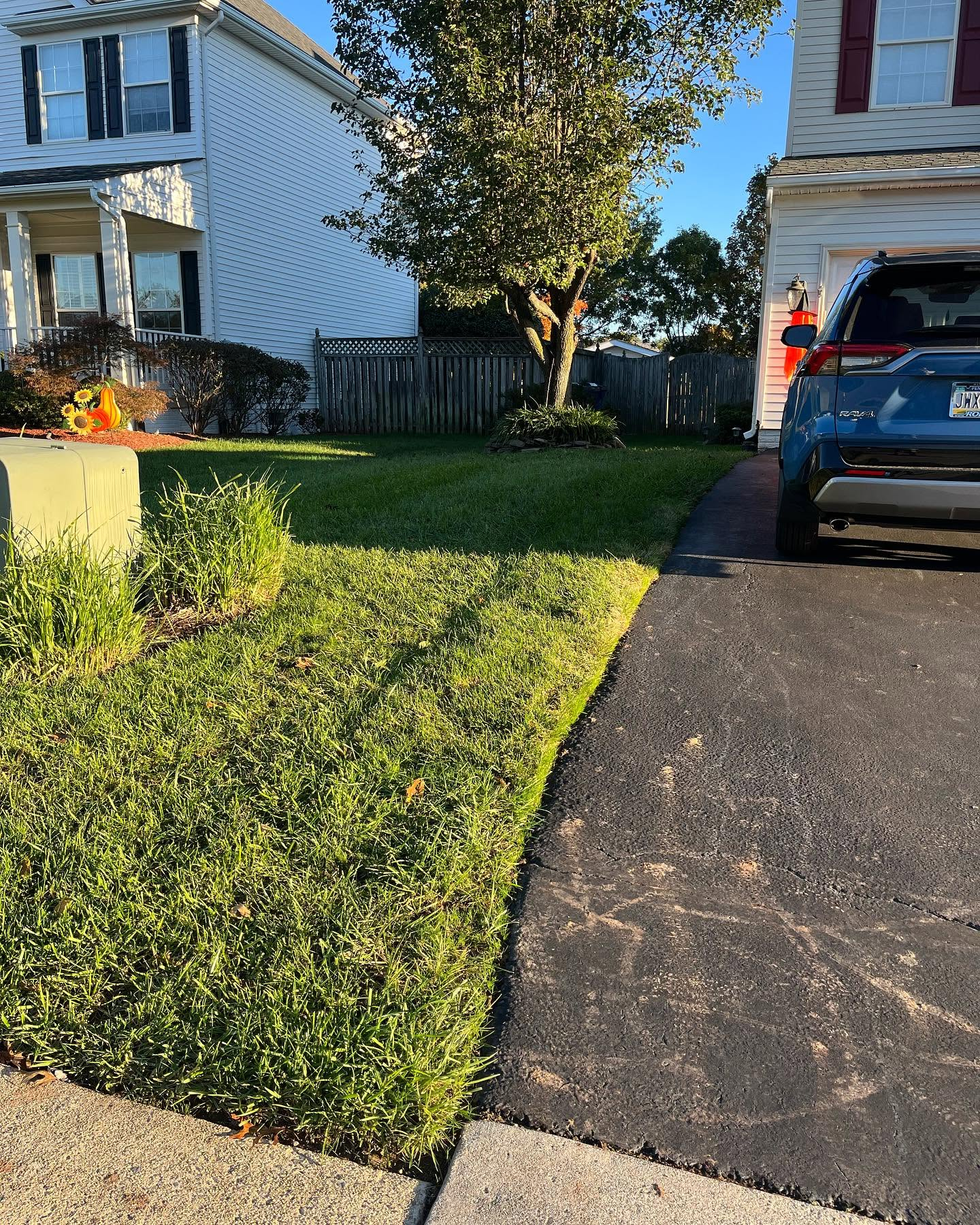 Suburban house driveway with a blue SUV, green lawn, and trees in front of a wooden fence.