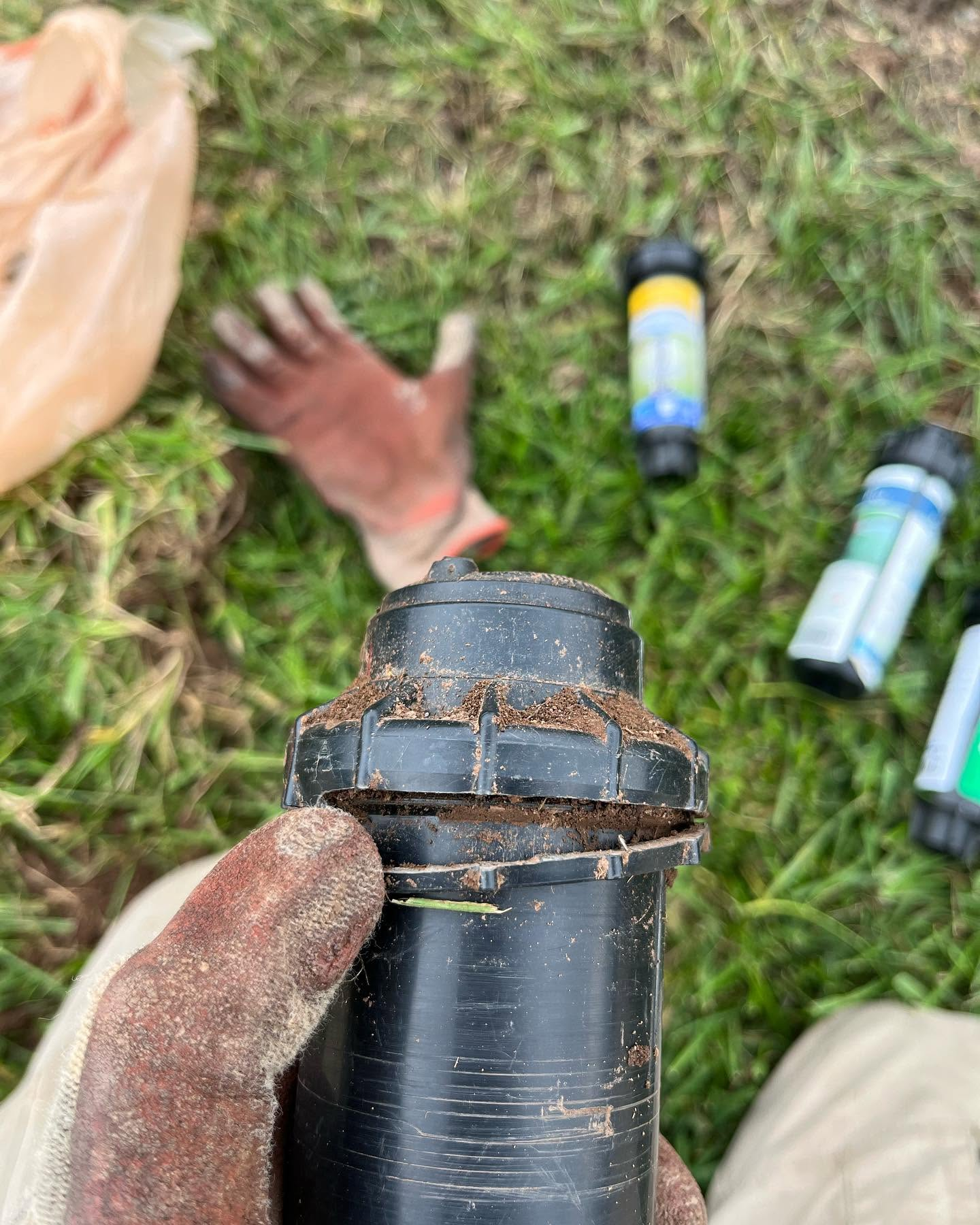 Close-up of a dirty black pipe fitting in grass, with a gloved hand and small bottles in the background