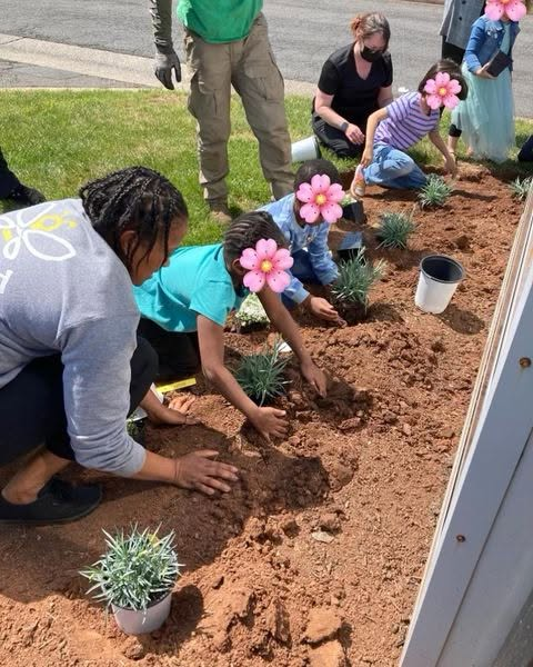 People planting flowers in a garden bed beside a building.