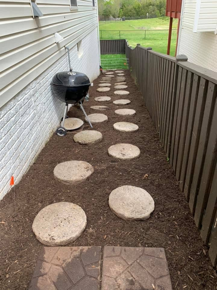 Narrow backyard path with round stepping stones, mulch, and a small grill beside the house