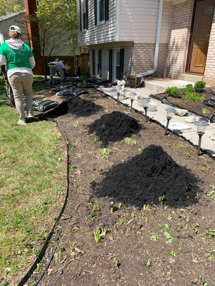 Mulch beds along a house foundation, with a person standing by a garden hose in the yard.