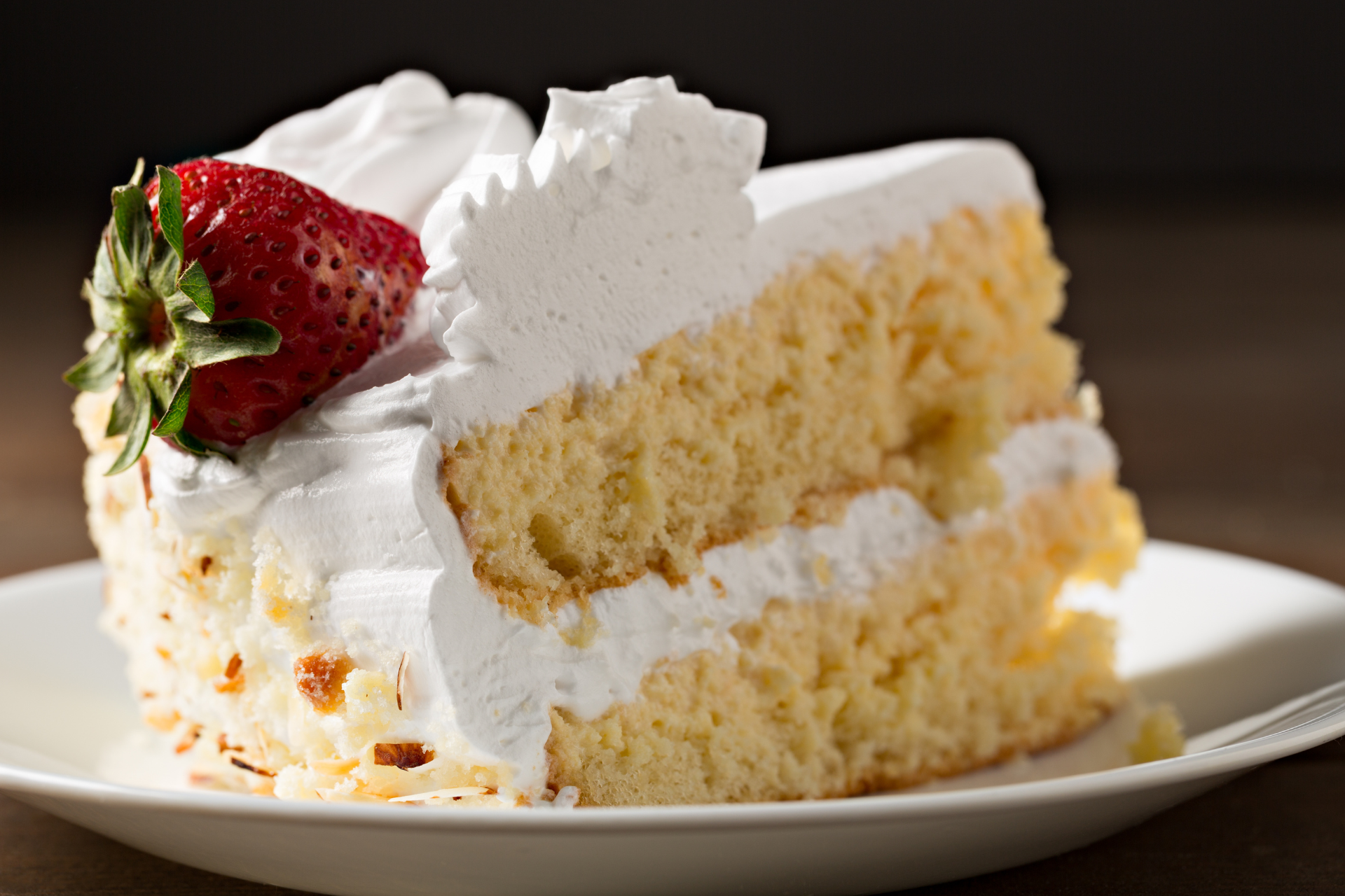 A slice of layered yellow cake topped with white frosting and a whole fresh strawberry on a white plate.