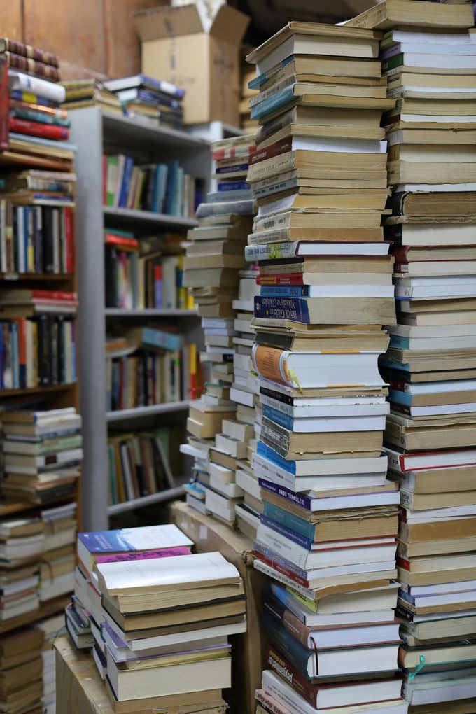 Stacks of books piled high in a cramped bookstore aisle, with shelves of books in the background