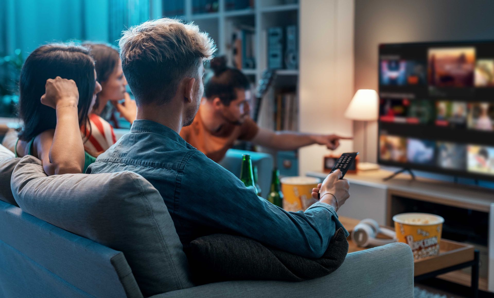 Friends on a couch watch TV, one holding a remote, another points. Snacks and drinks are nearby.