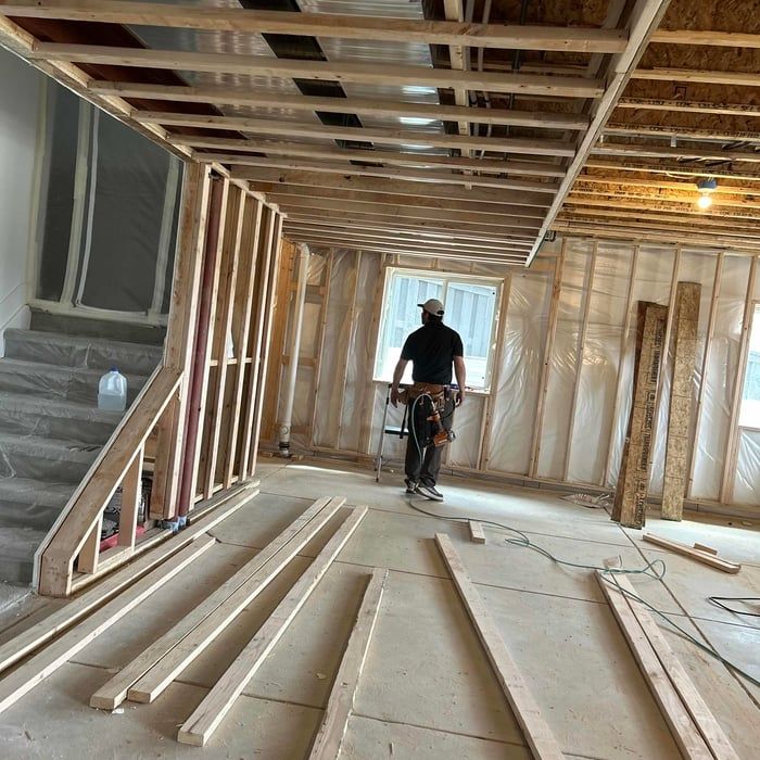 Interior of a house under construction. Carpenter standing near window, framed walls, exposed ceiling beams, wood planks on floor.