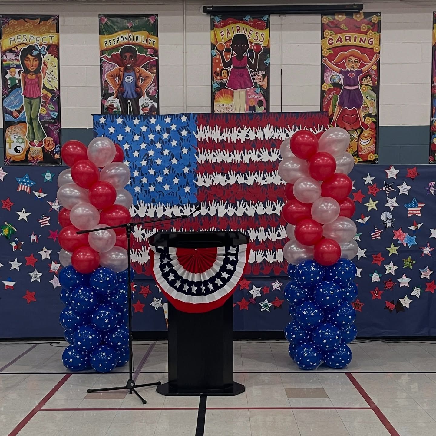 American flag backdrop with podium, flanked by red, white, and blue balloon columns; patriotic setting.