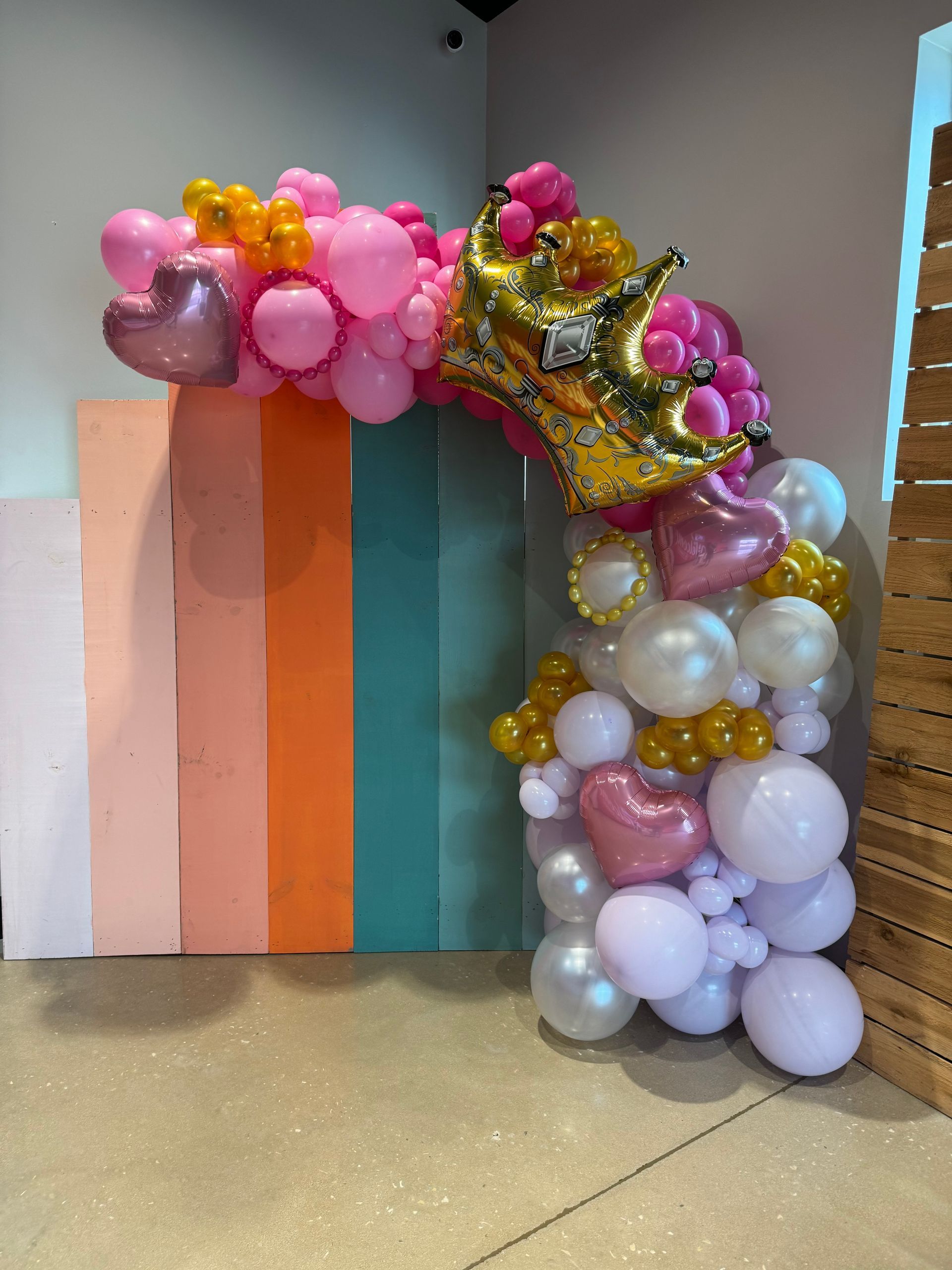 Pink, gold, and white balloon arch with a gold crown and heart balloons against a pastel backdrop.