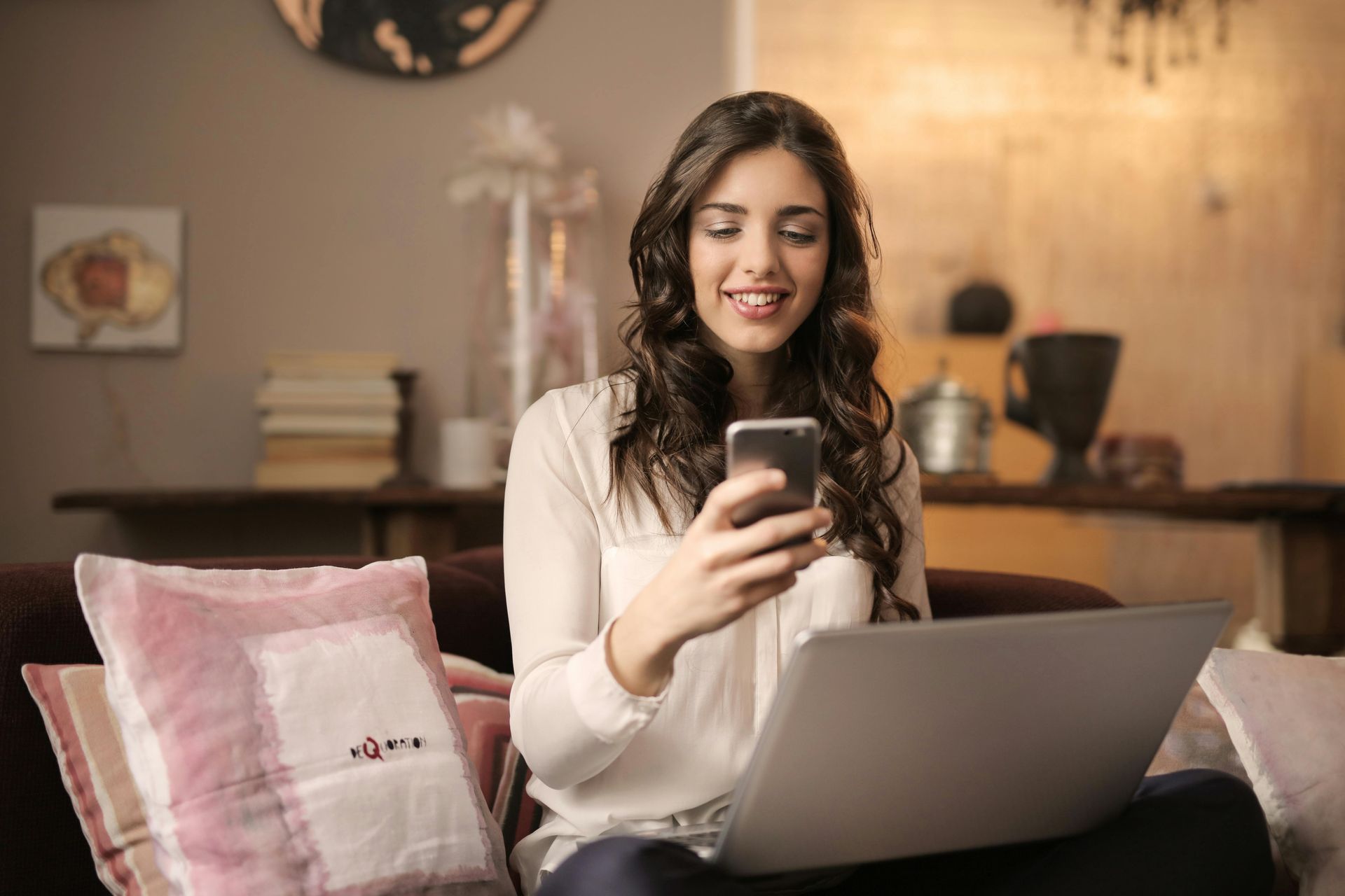 Woman with laptop and phone smiles while sitting on a couch. Living room setting, neutral colors.