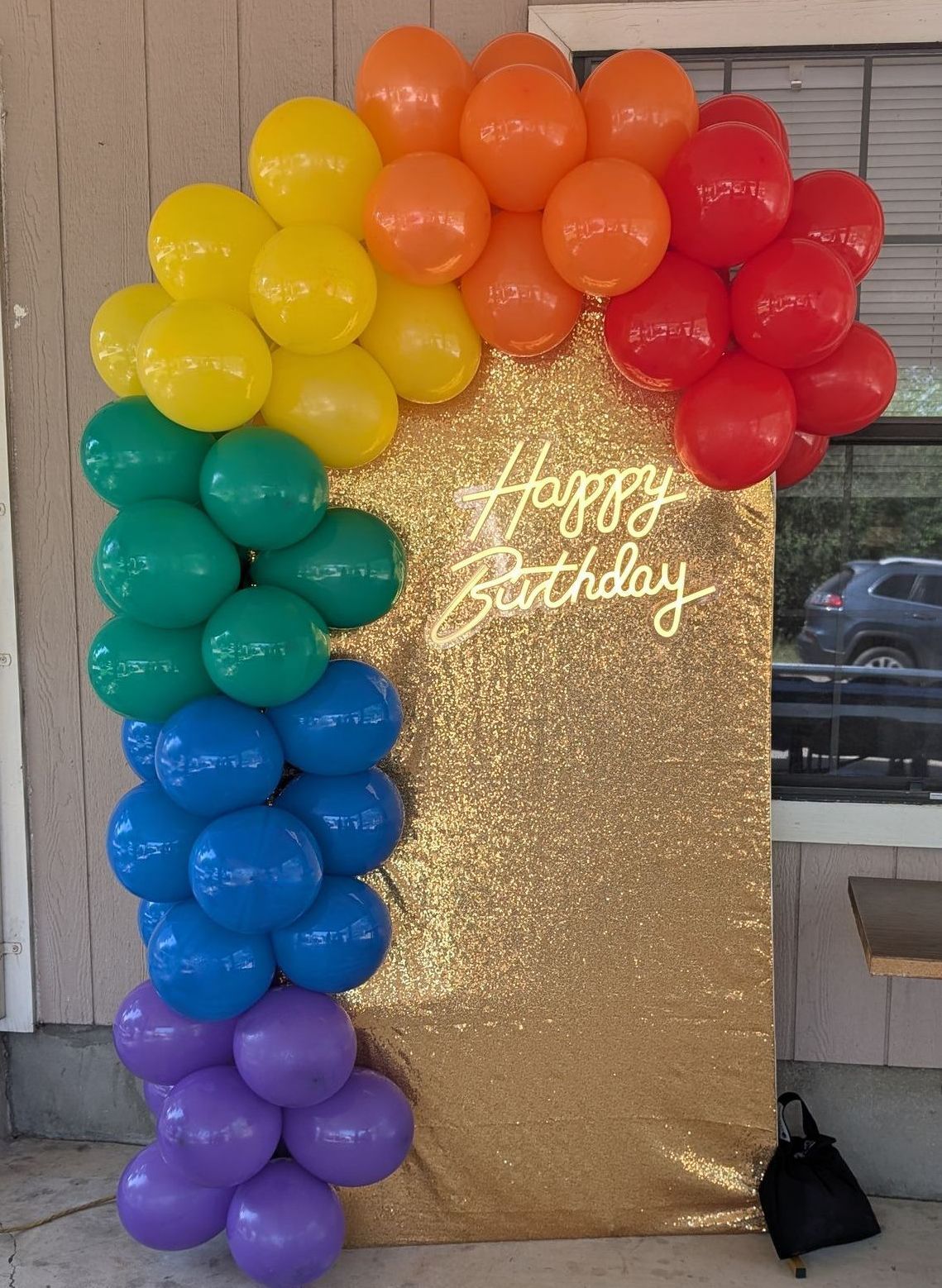 Rainbow balloon arch with a gold sequined backdrop that says