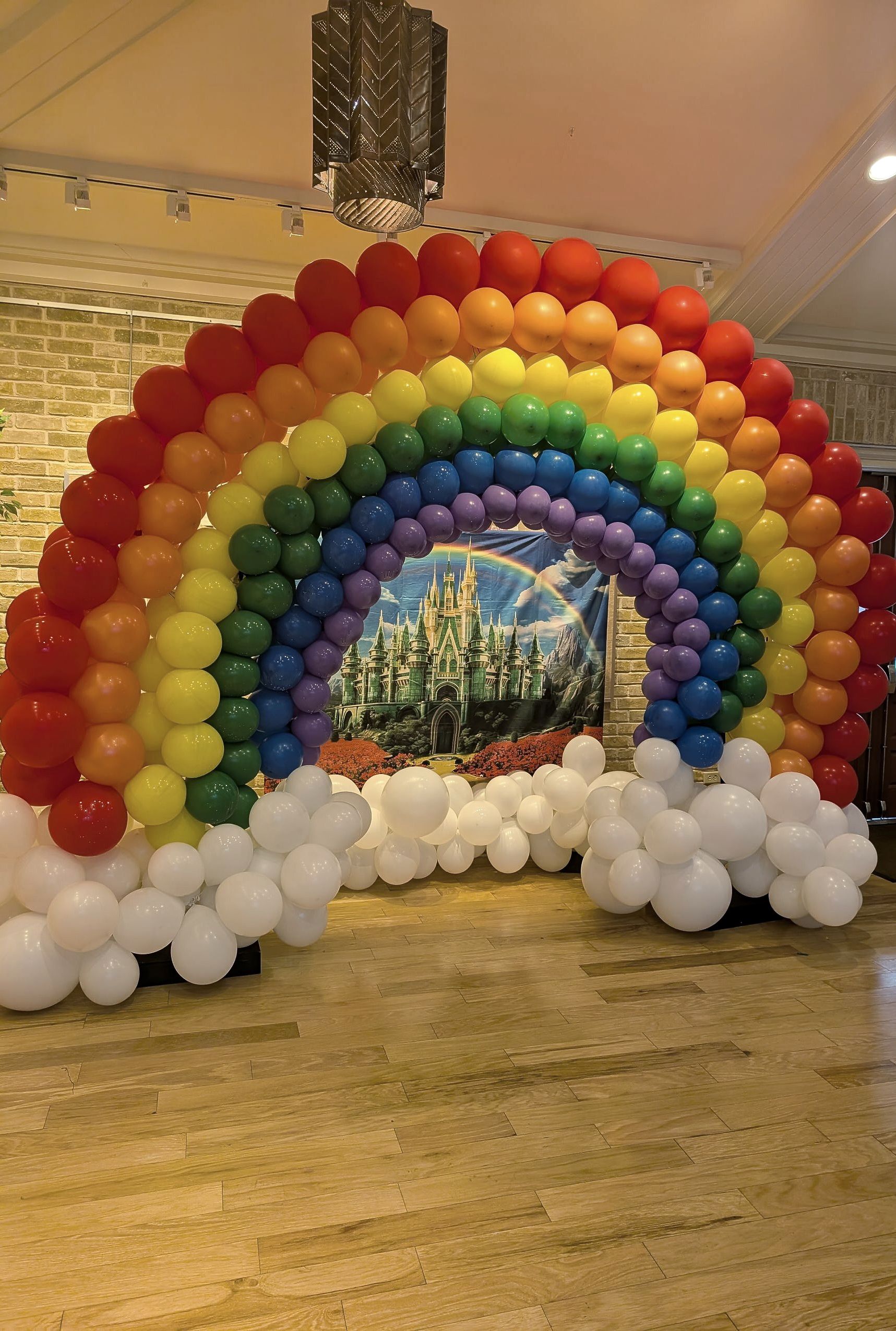 Rainbow balloon archway with white cloud balloons, in front of a castle painting.