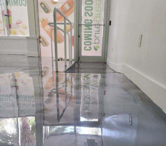 A smooth, high-gloss grey metallic epoxy floor reflecting a storefront entrance with white walls and a metal railing.