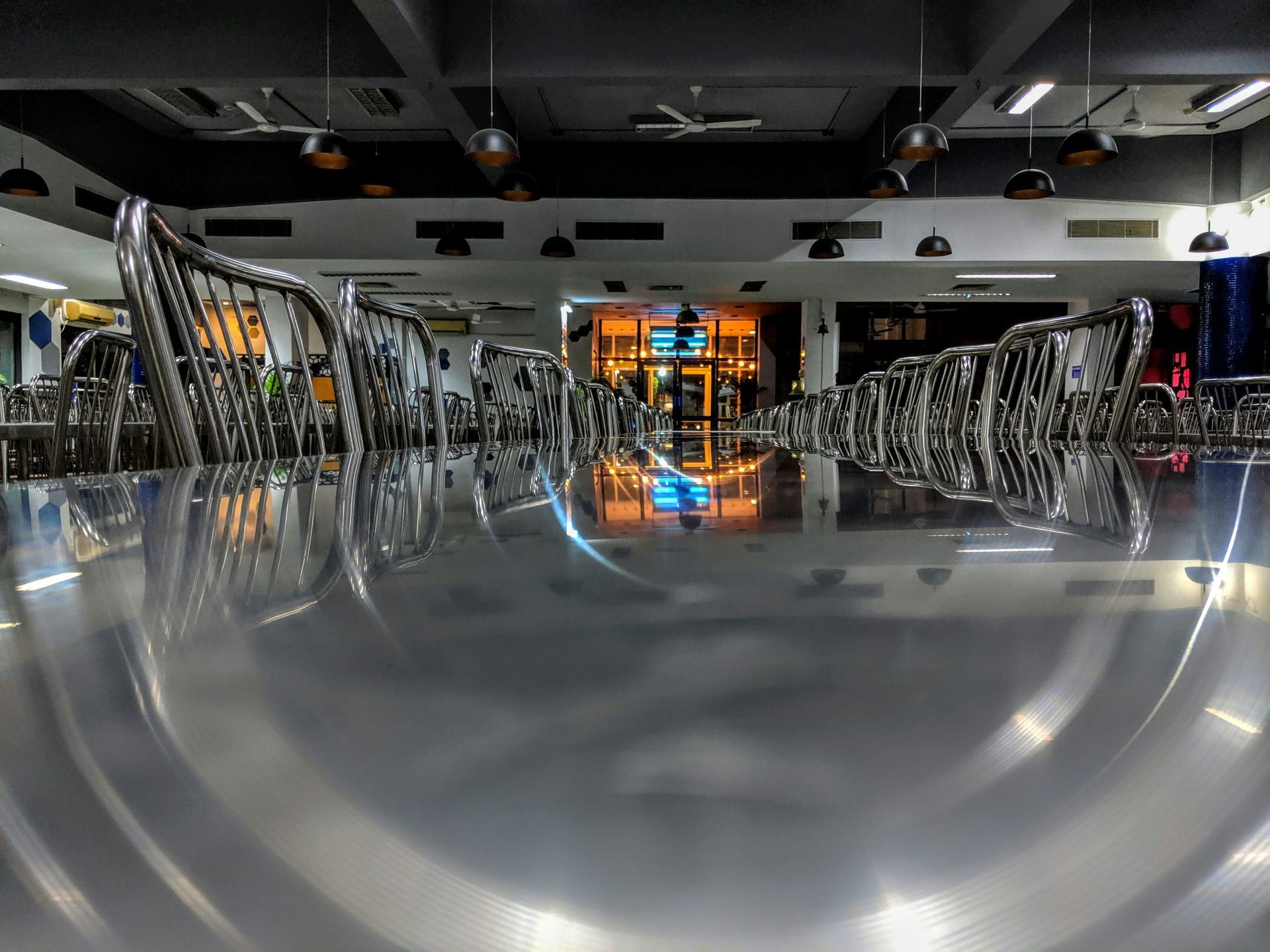 A low-angle view across a polished, reflective metal dining table, looking toward rows of empty metal chairs in a hall.