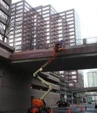 Workers in a bucket lift repairing a bridge over a city street, with high-rise buildings in the background
