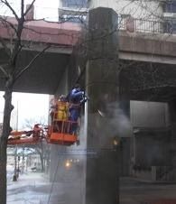 Workers in an orange lift cleaning a concrete bridge column with spray mist rising
