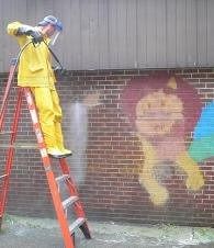 Person on a ladder sprays a brick wall with paint over colorful graffiti.