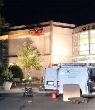 Nighttime scene of a lit building exterior with a utility van, cones, and equipment parked out front
