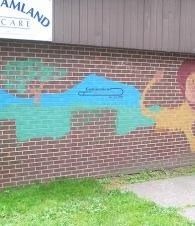 Brick wall with colorful mural of a blue animal and yellow flower beneath a sign reading “MAMLAND CARE”
