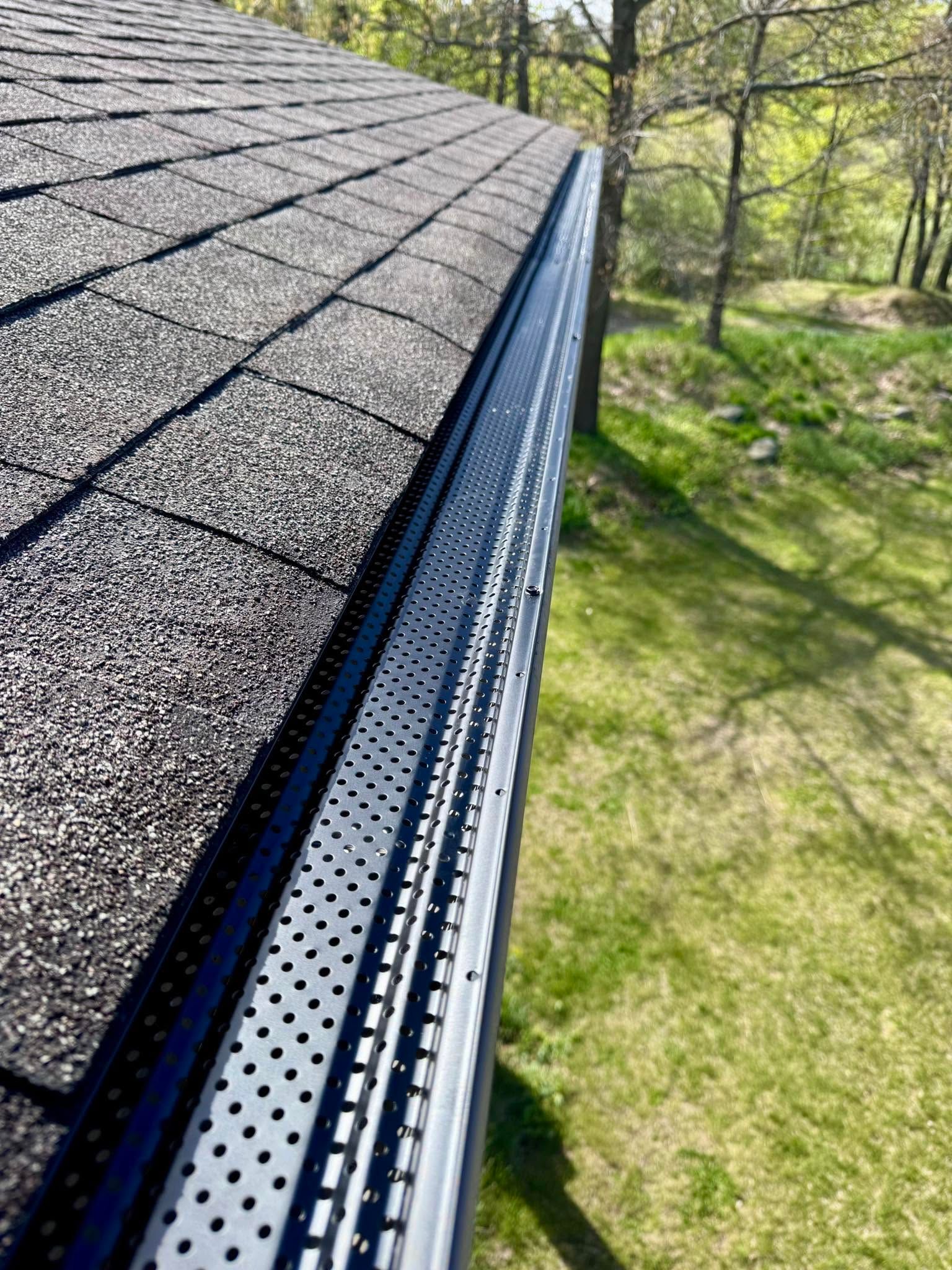 Roof edge with gray shingles and gutter beside a grassy yard with trees.