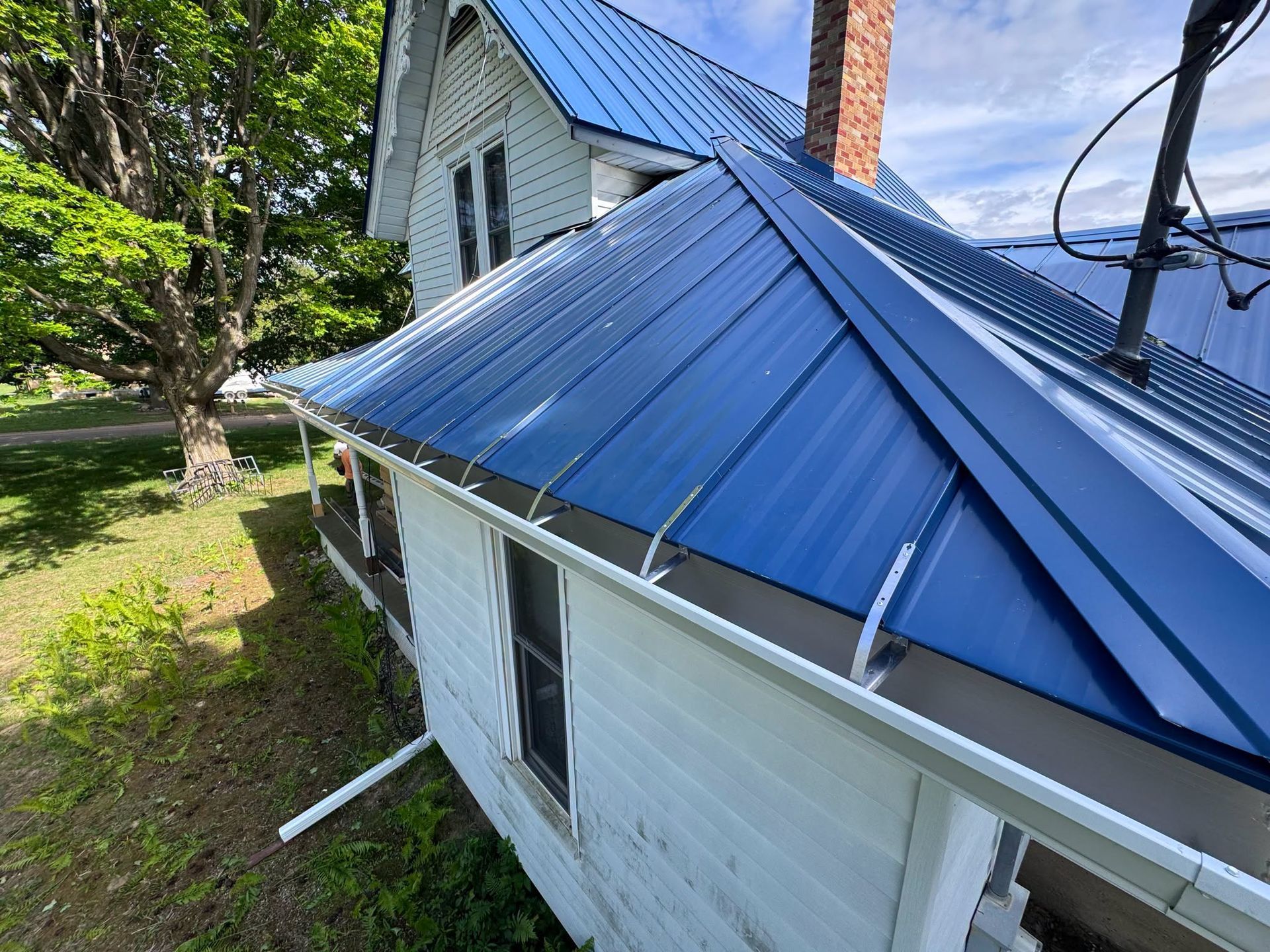 White house with a blue metal roof and chimney, viewed from above amid trees and yard.